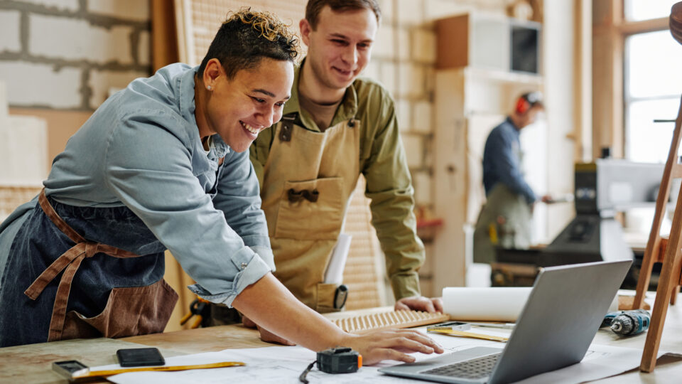 Warm toned portrait of animated carpenters using laptop and discussing furniture designs in workshop